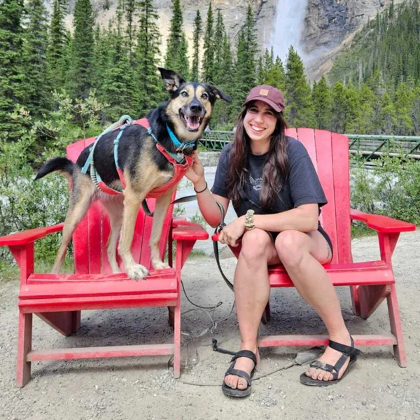 Woman sits on a red Adirondack chair beside a cheerful dog wearing a harness outdoors near a waterfall and trees in a park setting.